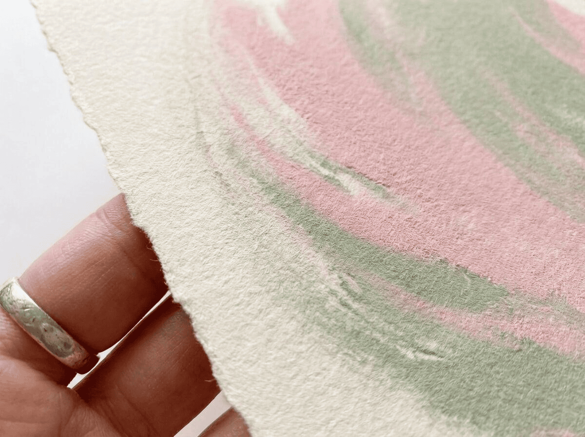 A macro shot showing the detail of the archival fine art paper. The viewer can see the subtle grain of the paper and the rich, velvety quality of the pastel pink and green pigments. A hand is seen lightly touching the edge of the print to signify quality and texture.